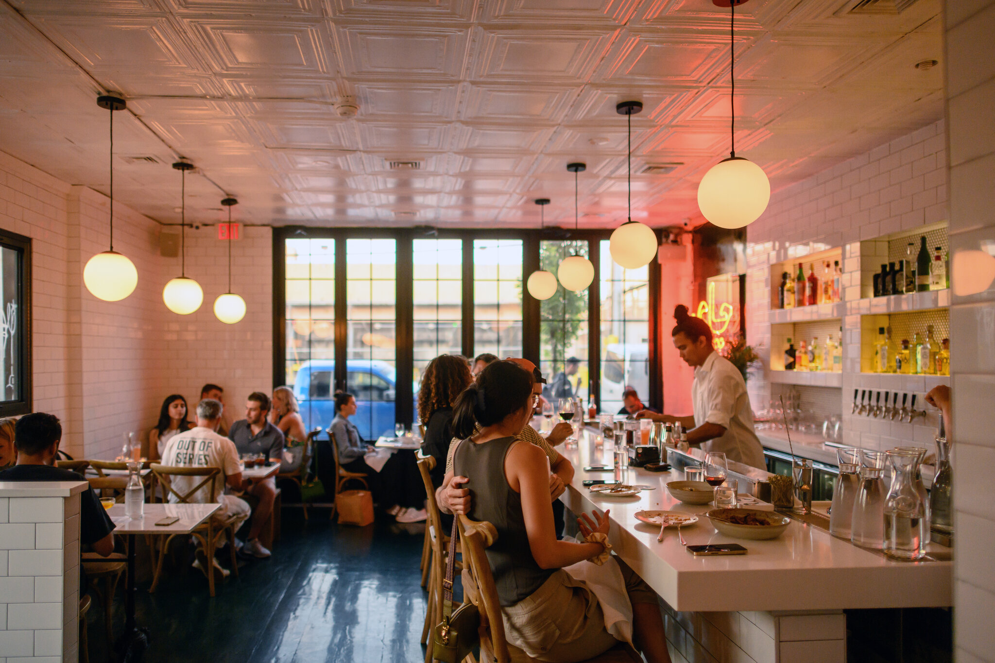 Lyst restaurantinteriør med gjester ved bardisk og bord, store vinduer og varme lamper.